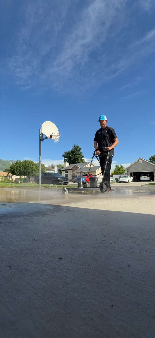 Pure Exterior crew member running a commercial hot-water surface cleaner on a residential driveway in Farmington, Utah, with visible steam and clean concrete behind the machine