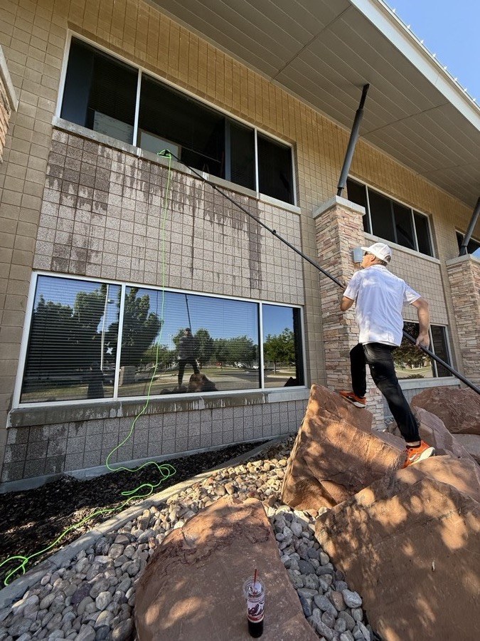 Pure Exterior team member cleaning the exterior windows of a tan-tile commercial office building in Sandy, Utah, using a water-fed pole and pure water system above a landscaped rock flowerbed